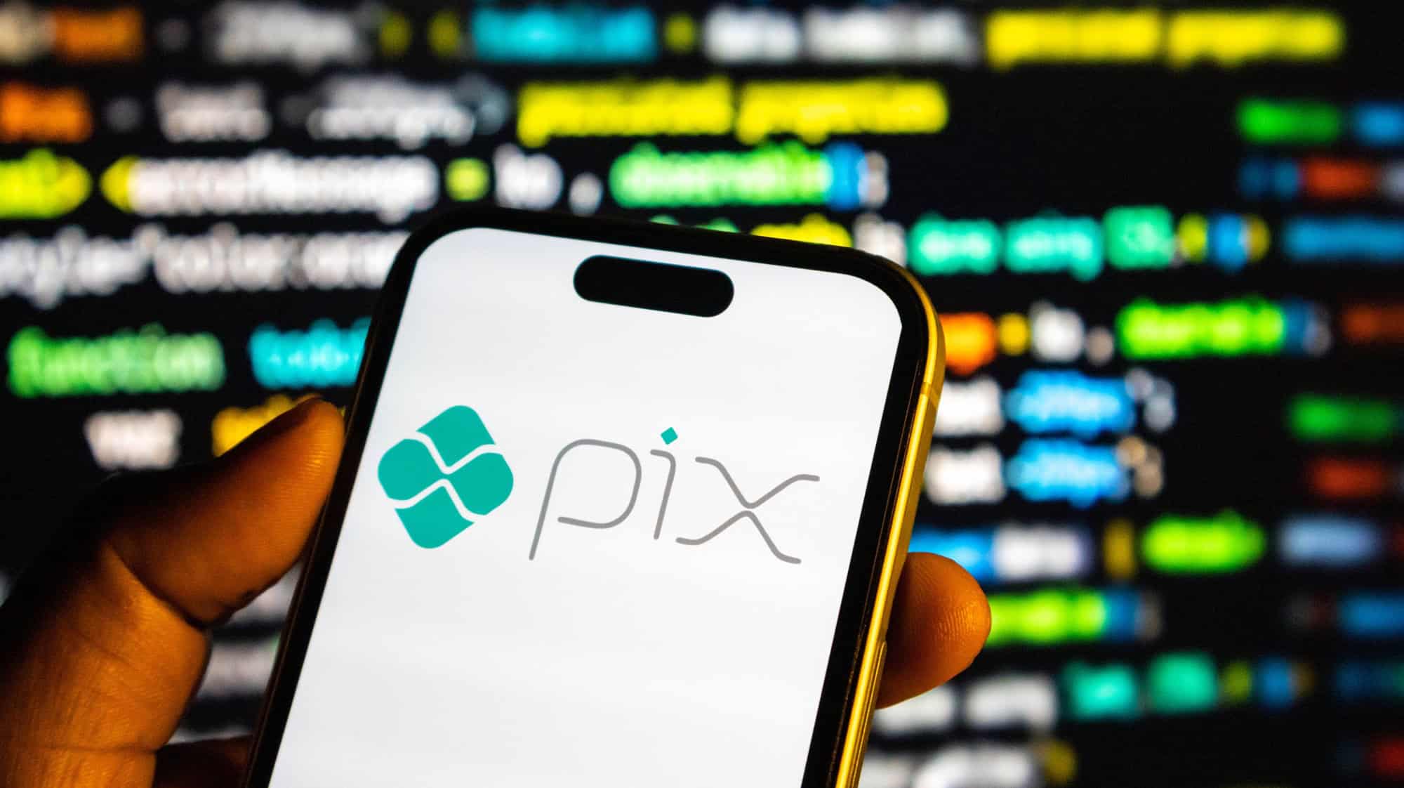 Pessoa segura um smartphone com a marca Pix na tela, diante de um fundo desfocado com informações e gráficos digitais.