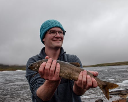 A man standing in a lake while wearing a bobble hat holding up a big fish with both hands